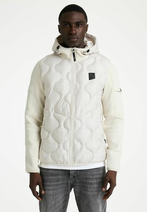 Man draagt een witte doorgestikte capuchonjas en een grijze spijkerbroek, staand tegen een effen witte achtergrond, kijkt recht vooruit.