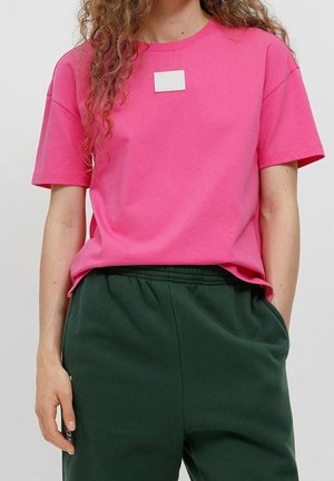 Różowa bawełniana koszulka z krótkim rękawem z kwadratową białą etykietą na klatce piersiowej. W połączeniu z ciemnozielonymi spodniami z elastycznym pasem.