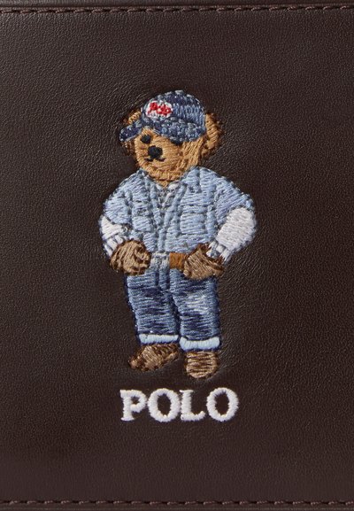 Κεντημένη αρκουδάκι που φοράει τζιν σύνολο και καπέλο, στέκεται με τα χέρια στη μέση πάνω από τη λέξη "POLO" σε σκούρο καφέ δέρμα.