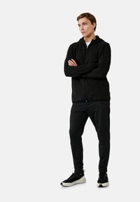 Zwarte zip-up hoodie en bijpassende broek van gladde stof, met een strakke pasvorm, lange mouwen en elastische boorden, gecombineerd met zwarte sneakers.