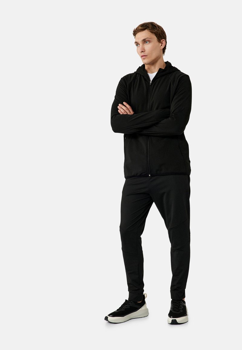 Zwarte zip-up hoodie en bijpassende broek van gladde stof, met een strakke pasvorm, lange mouwen en elastische boorden, gecombineerd met zwarte sneakers.