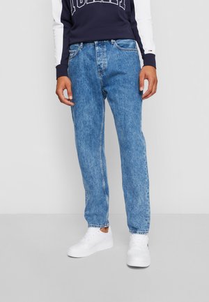 Homme portant un jean droit bleu, un sweat-shirt bleu marine avec des manches blanches et des baskets blanches, se tenant devant un fond uni.