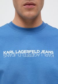 Zila kokvilnas T-kreklu ar apaļu kaklu, ar baltu drukātu tekstu "KARL LAGERFELD JEANS" uz priekšpuses, rakstīts treknā, mūsdienīgā fontā.