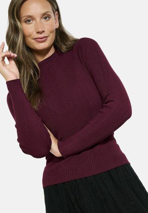 Pull en bordéau côtelé avec des manches longues et un col rond, présentant un design ajusté et un tissu texturé. Porté avec une jupe noire.