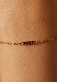 Gouden kettingarmband met drie rode edelstenen gezet in een ronde staaf, gelegen op een gladde, gebruinde huid. Eenvoudig ontwerp, fijne textuur.