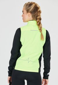 Neon groene jas met zwarte mouwen, ritssluiting aan de voorkant en een reflecterende accent aan de achterkant. Heeft een grafisch patroon van een hartslaglijn.