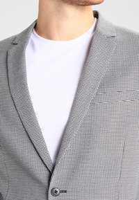 Mężczyzna w czarno-białym żakiecie w kratę houndstooth, noszący prostą białą koszulkę z okrągłym dekoltem, odsłaniającą kołnierz i górną część klatki piersiowej.