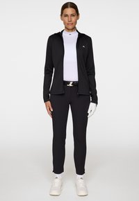 Kvinna iklädd svart zip-up jacka, vit höghalsskjorta, svarta byxor, vit golfhandske på höger hand, vita sneakers, stående mot vit bakgrund.