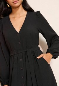 Čierne rebrované šaty s dlhými rukávmi, výstrihom do V, gombíkmi na prednej strane, opaskom v páse a bočnými vreckami. Jemná textúra s plynulou siluetou.