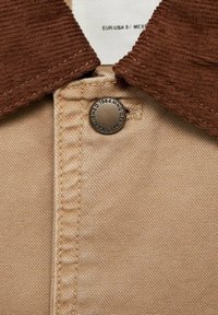 Beige fabric with a corduroy brown collar, featuring a metal button with engraved details. Visible stitching adds texture.