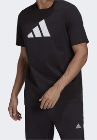 Muž oblečený v černém tričku a kalhotách Adidas s bílými logy, stojící proti jednobarevnému světlému pozadí.