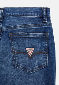 Tumesinine pesu denim teksad, pisut kulunud. Tagataskul on kolmnurkne punane ja valge logo lapik, millel on pruunid õmblused.