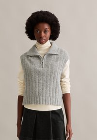 Hellgrauer, gerippter ärmelloser Pullover mit hohem Kragen und Reißverschluss, getragen über einem cremefarbenen Rollkragenpullover, kombiniert mit einem dunklen Rock.