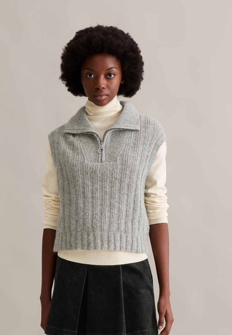 Hellgrauer, gerippter ärmelloser Pullover mit hohem Kragen und Reißverschluss, getragen über einem cremefarbenen Rollkragenpullover, kombiniert mit einem dunklen Rock.