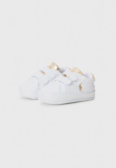 Chaussures synthétiques blanches pour tout-petits avec des bandes Velcro, des accents dorés sur la languette et un petit logo sur le côté. Semelle en caoutchouc plate.