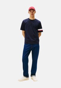 Camiseta de algodón azul marino con el logo "HILFIGER" en relieve, acentos de rayas rojas y blancas en las mangas, combinada con jeans azules y zapatillas blancas.