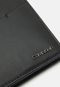 Black leather wallet with a textured finish, featuring a smooth panel and embossed "Calvin Klein" logo in silver. Subtle stitching details.