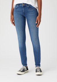 Jeans de mezclilla azul ajustados con una cintura de media altura, diseño de cinco bolsillos y un ligero desvanecimiento. Combinados con zapatillas de lona negras.