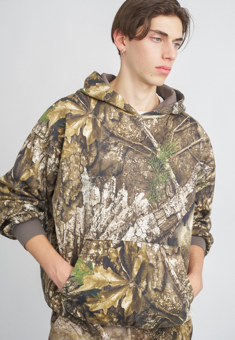 Camo hoodie featuring a leaf and bark pattern in browns and greens, with a front pocket and drawstring hood, placed against a gray backdrop.