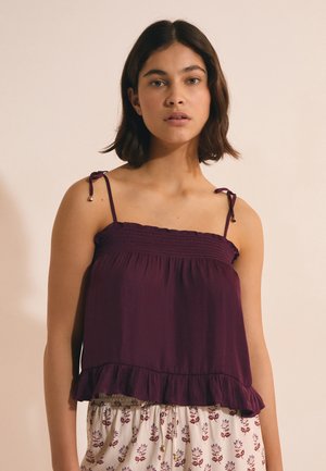 Młoda kobieta o ciemnych włosach do ramion, ubrana w fioletowy top bez rękawów z wiązanymi ramiączkami oraz białe spodnie w kwiatowy wzór.