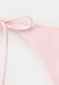 Bas de bikini rose clair avec un nœud noué sur le côté sur un fond blanc uni, montrant la texture du tissu et les détails des coutures.