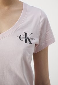Svetle růžové bavlněné tričko s výstřihem do V a krátkými rukávy. Na hrudi se nachází černé logo "CK" a text "Calvin Klein Jeans".