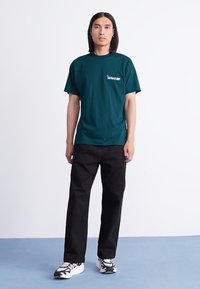 Mann mit langen dunklen Haaren, der ein dunkelgrünes T-Shirt, schwarze Hosen und weiße Sneakers trägt, steht auf einem blauen Boden vor einer weißen Wand.