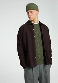 Tamsiai rudas marškinėlių švarkas su tekstūruotu audiniu ir sagų uždarymu; apsivilktas ant žalio kabelinio megztinio. Modelis dėvi derančią žalią kepurę.