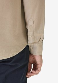 Beige button-up paita, jossa on pitkät hihat, terävä kuuppa ja hienovarainen rakenne, jotka on pistetty tummiin housuihin. Käsi näkyy sivussa.