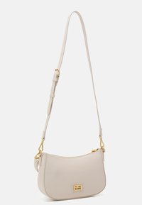 Beżowa torba na ramię o gładkiej fakturze, owalnym kształcie, złotych akcesoriach, regulowanym pasku i małej plakietce z logo z przodu.