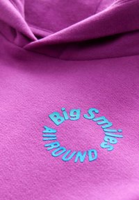 Lila Kapuzenpullover mit gesticktem blauen Text, der "Big Smiles All Around" liest, mit weicher Textur und lässiger Form.