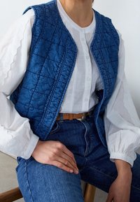 Quiltad blå väst i ett glänsande material, bärs över en vit blus med puffärmar, kombinerad med blå jeans och ett brunt bälte.