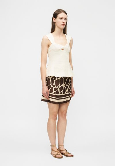 Γυναίκα με αμάνικο λευκό τοπ και καφέ φούστα με σχέδιο καμηλοπάρδαλη, στέκεται με τα χέρια χαλαρά στο πλάι, πάνω σε λευκό φόντο.