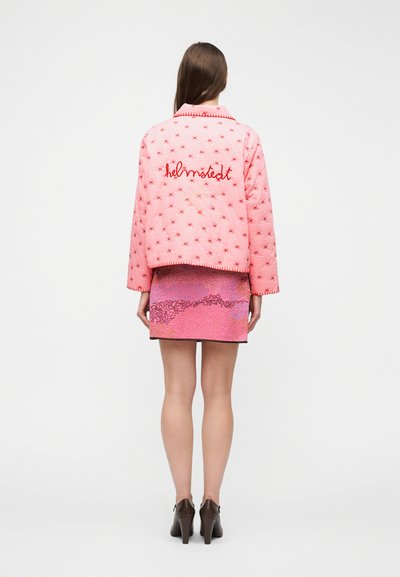 Γυναίκα με ροζ μπουφάν καπιτονέ με κόκκινα αστέρια και το κέντημα "helmsledt" στην πλάτη, συνδυασμένο με ροζ υφασμάτινη μίνι φούστα και καφέ ψηλοτάκουνα.