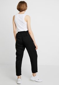 Osoba stojící zády, oblečená v bílé bezrukávové topu, černých kalhotách s vysokým pasem a bílých teniskách na jednobarevném pozadí.