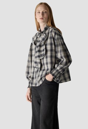 Frau mit langen roten Haaren, die eine schwarz-weiß karierte Bluse mit großer Schleife und schwarze Hose trägt, steht mit einer Hand in der Tasche.