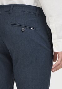 Marinblå byxor i texturerad tyg, med ett bakfickor med knapp och en liten randig detalj nära fickan.