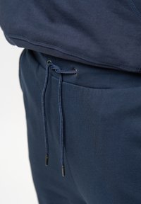 Mörkblå joggers med dragsko i midjan, har en slät textur, metallspetsar och en avslappnad passform. Synlig sömnadsdetalj.