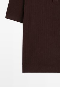 Chemise à manches courtes en tricot côtelé marron foncé avec une texture visible, présentée sur un fond blanc.
