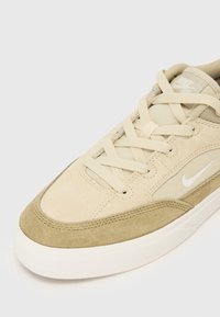 Bežinės zomšinės ir tinklinės sportinės bateliai su tamsiai žaliu viršeliu, plokščiomis raišteliais ir balta swoosh logotipu. Apvalus pirštas ir paminkštinta padas.