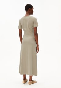 Sagegrünes Maxikleid aus weichem Stoff, mit kurzen Ärmeln, geradem Schnitt und hohem Kragen. Getragen mit beigen Slippers.