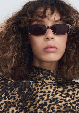 Ochelari de soare rectangularei închiși, cu rame groase, purtați de un model cu păr creț, asortați cu un top în stil leopard cu gât înalt.