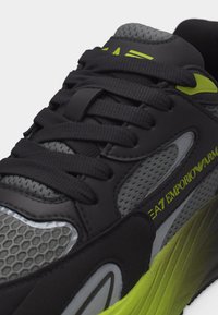 Κοντινή λήψη μαύρου και γκρι αθλητικού παπουτσιού με έντονες πράσινες λεπτομέρειες, πάνελ από δίχτυ και τη σήμανση "EA7 Emporio Armani" στο πλάι.