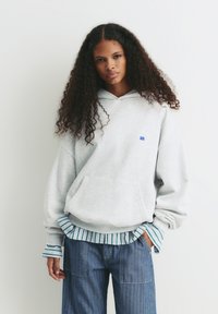 Felpa grigia chiara con tasca frontale, logo ricamato blu, indossata sopra una maglietta a righe blu e verdi. Jeans a gamba larga blu scuro.