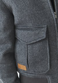 Giacca in misto lana grigia con taschino sul petto sinistro, polsini a costine e patch in pelle con logo. La cerniera è metallica e funzionale.