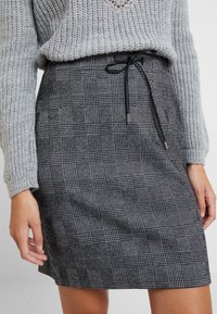 Maglione grigio a maglia indossato sopra una gonna a scacchi bianca e nera, con cintura regolabile e motivo testurizzato.