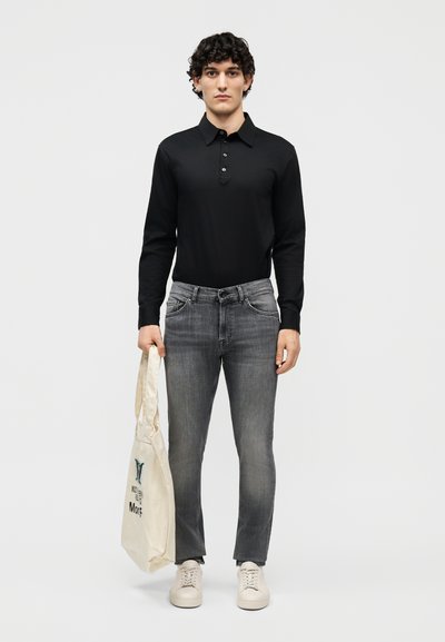 Bărbat purtând un tricou polo cu mâneci lungi negru, blugi gri skinny, adidași albi, ținând o geantă tote bej cu text negru și verde.