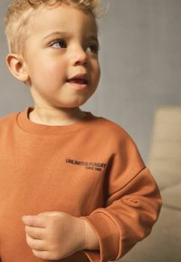 Sweatshirt orange avec le texte "UNLIMITED FUNDAY" et un col rond, avec des poignets côtelés et une texture douce. La main d'un enfant est partiellement visible.