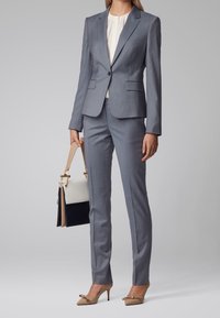 Grijs op maat gemaakt pak met een aansluitende blazer en slanke broek. De outfit is gecombineerd met een crèmekleurige blouse en beige hoge hakken, en houdt een handtas in twee tinten vast.