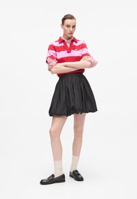 Rosa og rødstripet polo-skjorte med krage, kombinert med et svart, voluminøst skjørt. Svart loafers og kremfargede sokker fullfører antrekket.